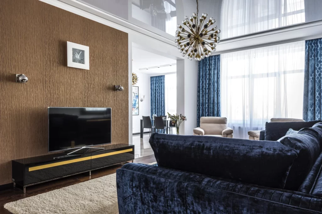 home Remodeling Elegant living room featuring a plush velvet sofa, textured accent wall, modern lighting fixture, and large windows with rich blue curtains.